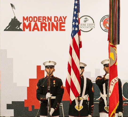 Three Marine honor guards in dress blues with rifles and flags in front of a 'Modern Day Marine' backdrop.